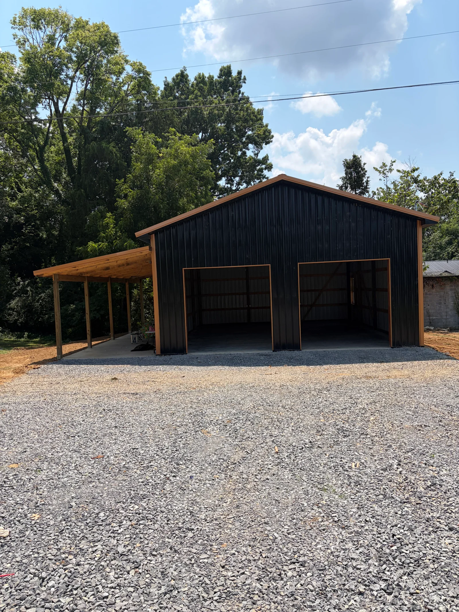 Black metal garage with wood trim, two large glass doors, and attached wood carport on gravel driveway