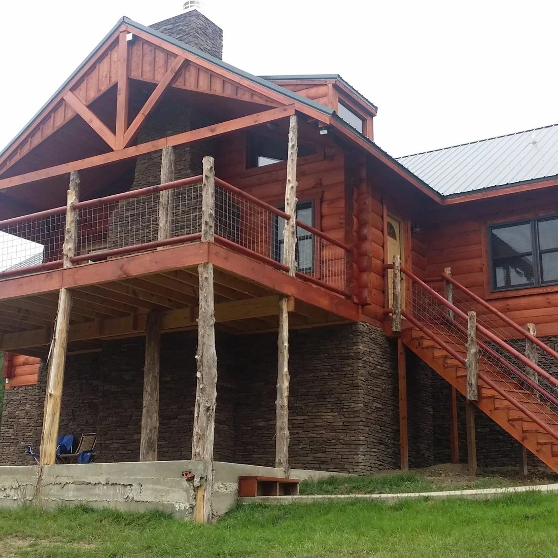 Red log home with stone foundation, timber deck framing, and cable railings